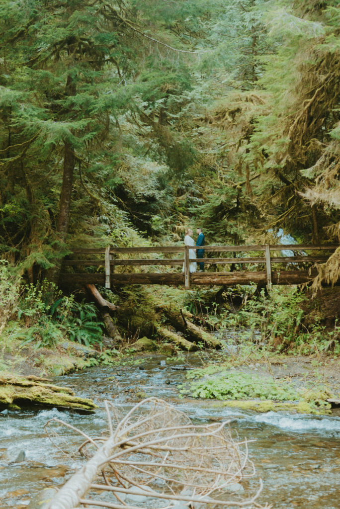 couple standing in the middle of a bridge in Olympic National Park forest, captured by destination elopement videographer, Alesia Films