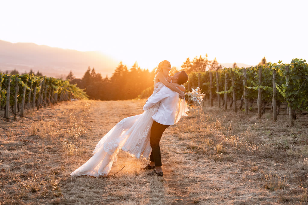 Portrait of groom carrying bride laughing during golden hour at the Domaine de Broglie vineyard after their winery summer elopement in Dayton, Oregon