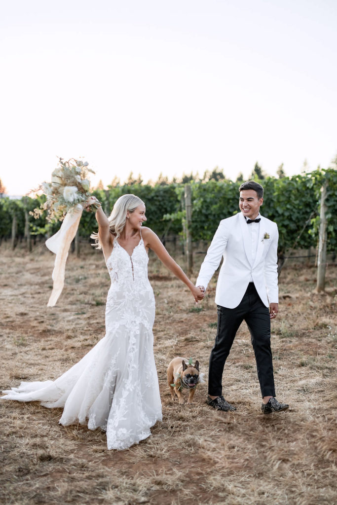 Newlyweds with their dog walking through Domaine de Broglie winery and vineyard and holding hands after their summer elopement in Dayton, Oregon