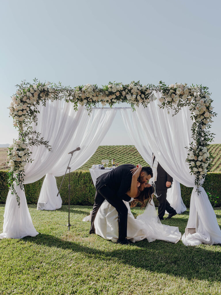 Groom dipping bride for romantic first kiss under chuppah at Temecula wedding ceremony