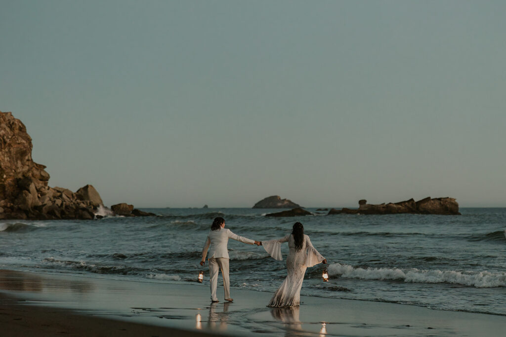 Brides frolicking in the waves holding lanterns after redwoods California wedding