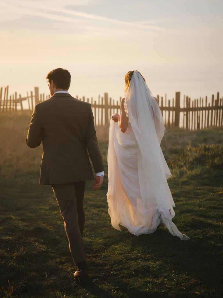 Bride and groom walking toward the shoreline as golden light casts soft shadows