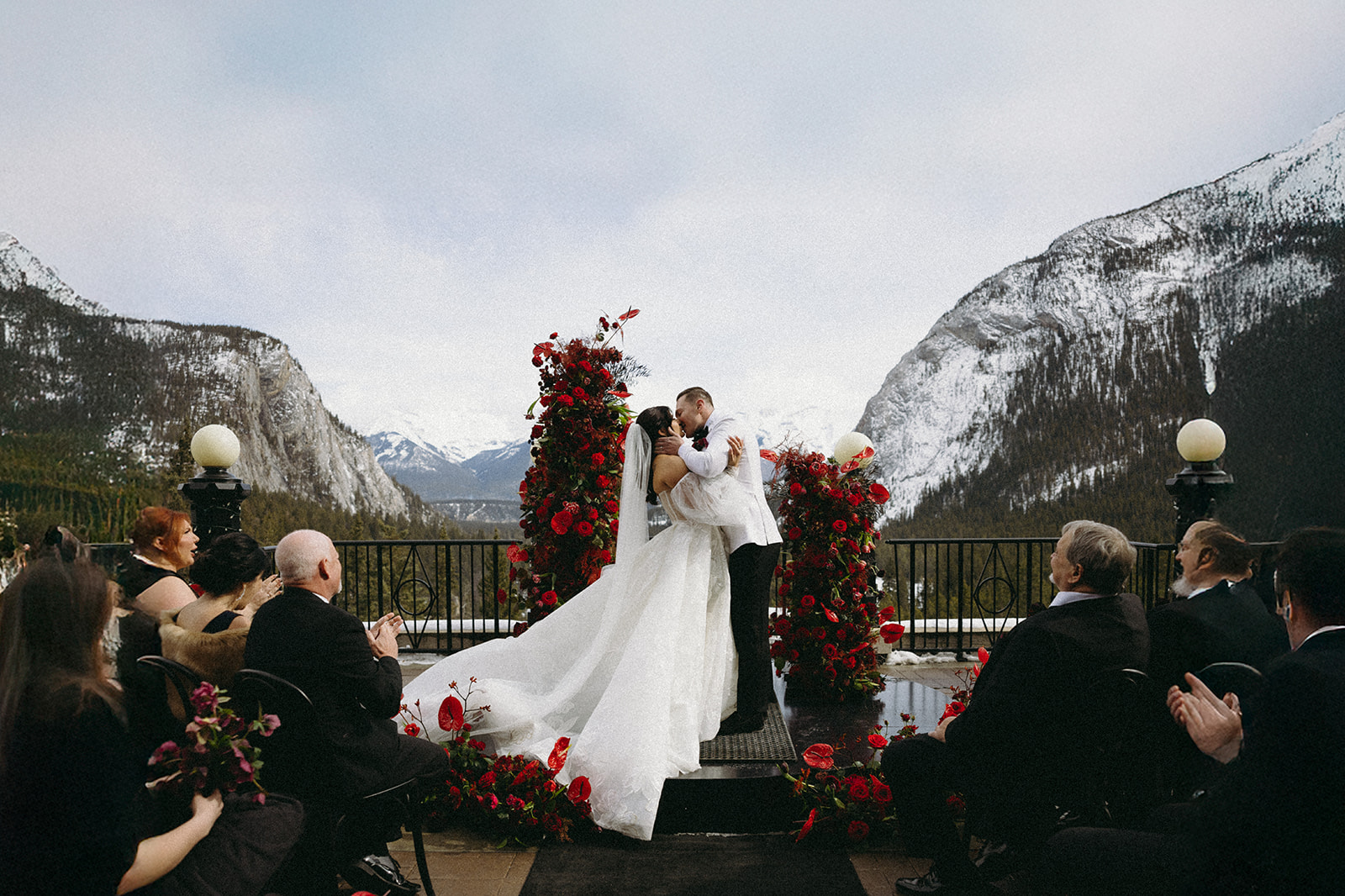 Couple kisses at the altar surrounded by red florals and snowy mountain views