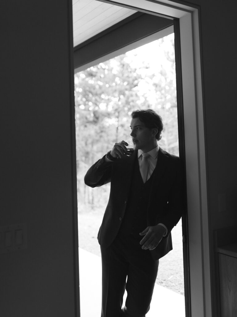Groom sipping a drink while standing in a doorway, dressed in a dark suit with trees in the background.