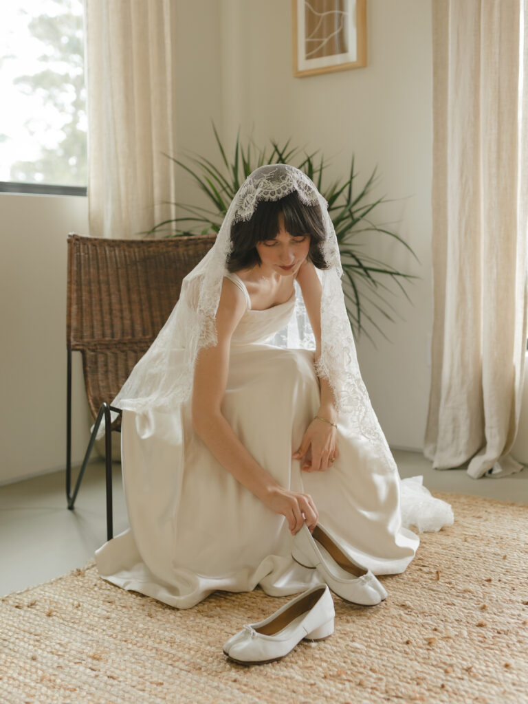 Bride in a lace veil slipping on white shoes while getting ready inside a softly lit room.