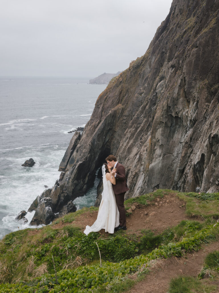 Groom kisses bride on the cliff during vintage-inspired Oregon Coast elopement.