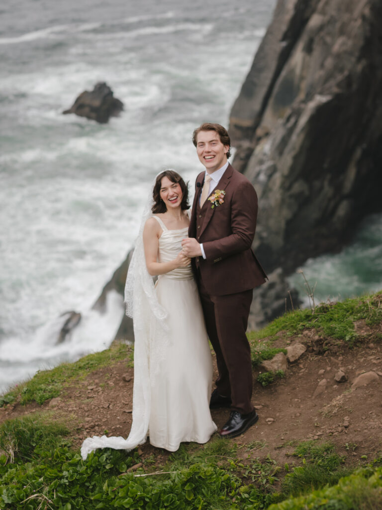 Couple laughing joyfully after elopement ceremony on the edge of a coastal cliff.
