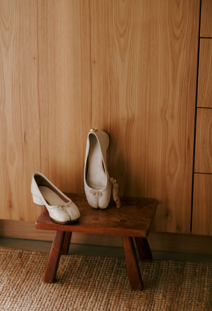 White wedding shoes with a gold ring on a rustic wooden stool in front of a light wood cabinet.