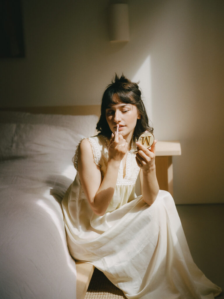 Bride applies lipstick by the window during a vintage-inspired Oregon Coast elopement.