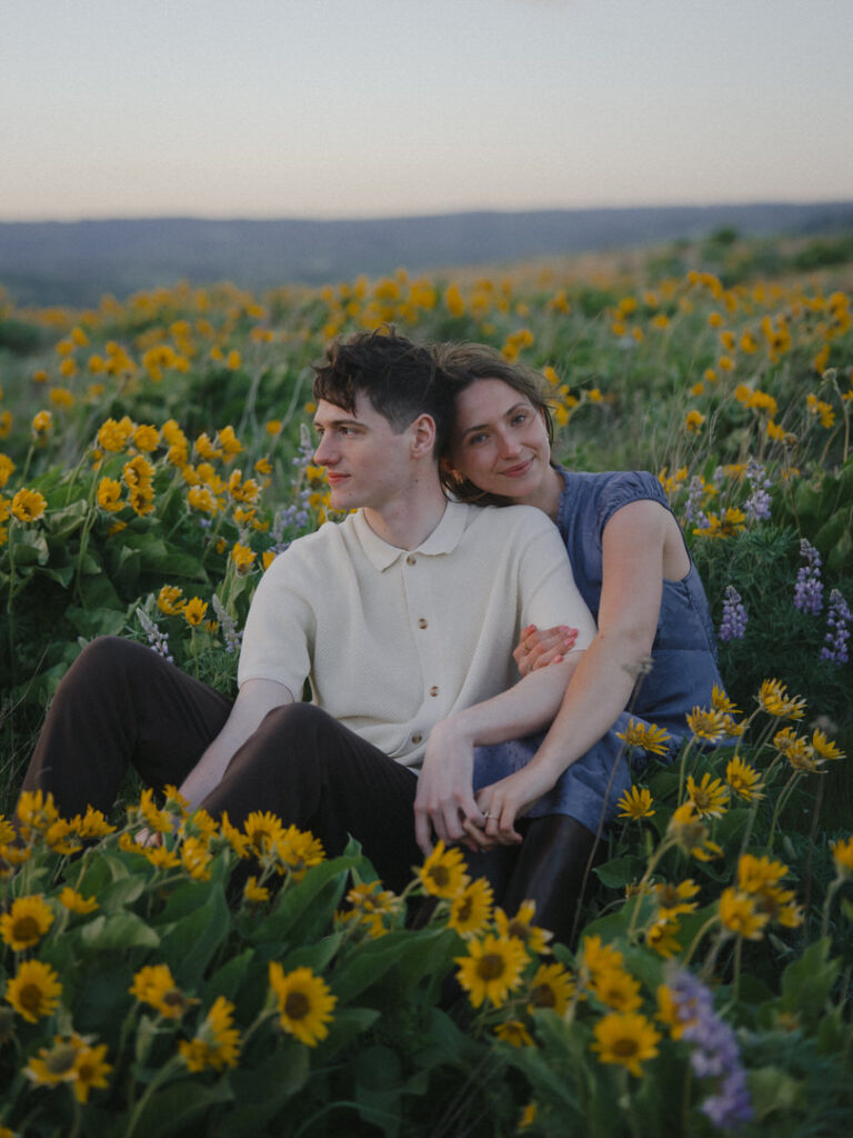 Couple sits close together in a hillside meadow filled with wildflowers, smiling as the day fades.