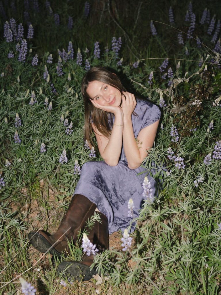 Woman in purple dress posing playfully in a patch of wild lupine flowers.