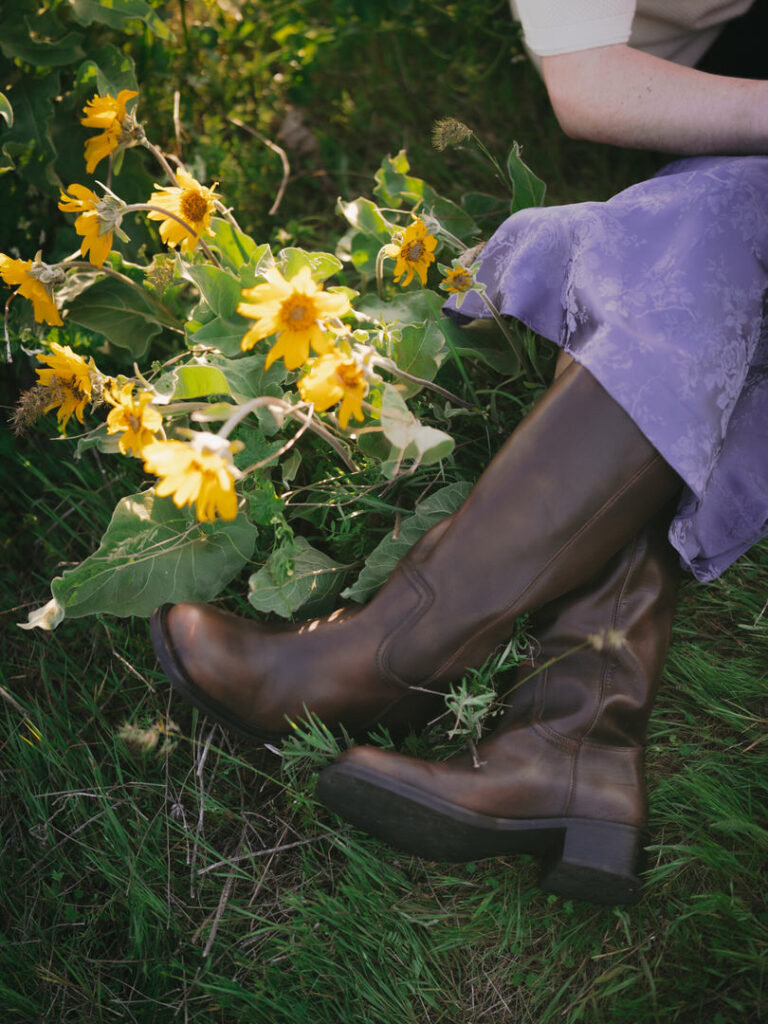 Close-up of tall brown boots resting near yellow wildflowers and a lavender dress during a springtime session.