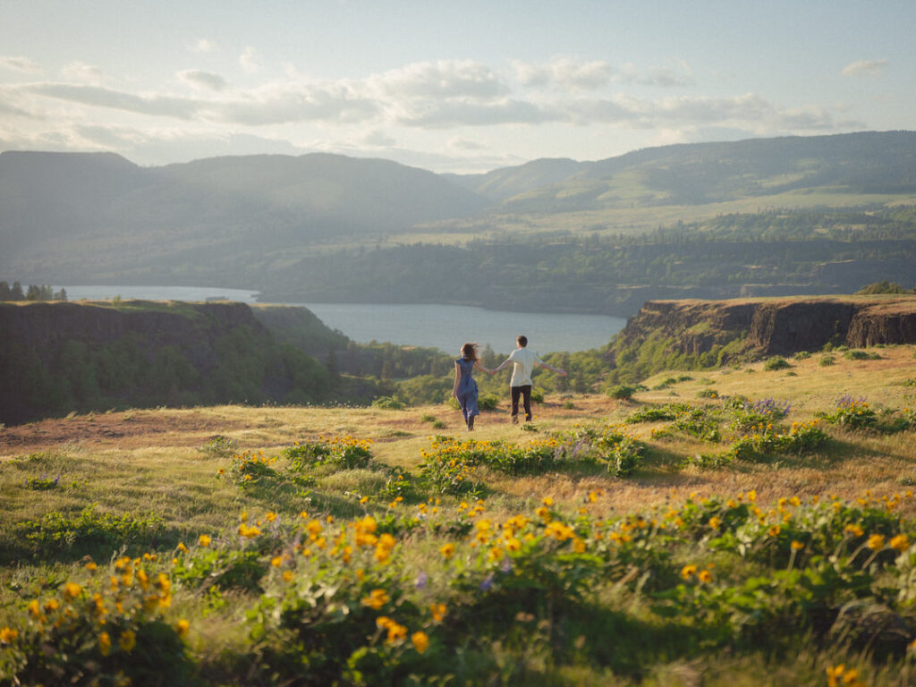 Couple holds hands walking through hills for their Rowena Crest wildflower engagement above the Columbia Gorge.