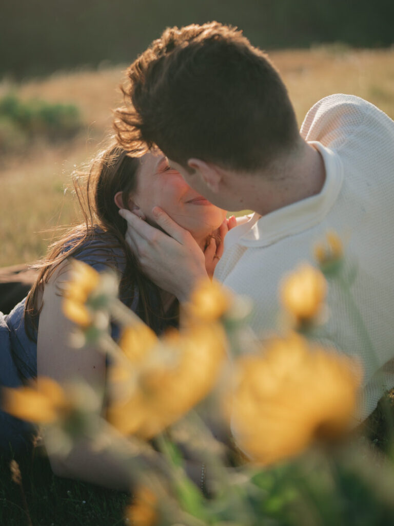Groom-to-be holds fiancée’s face as they laugh among yellow flowers in soft evening light.