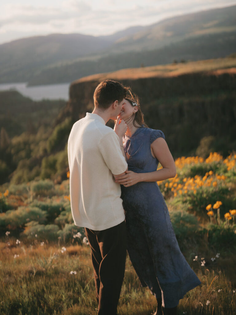 Couple shares a quiet kiss during golden hour, surrounded by vivid blooms and a scenic cliffside view.