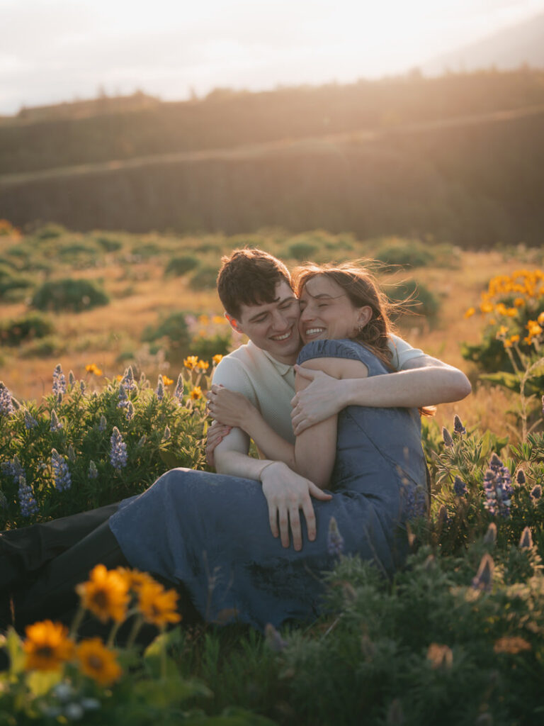 Laughing couple cuddles in a patch of lupine and balsamroot during their Rowena Crest wildflower engagement.