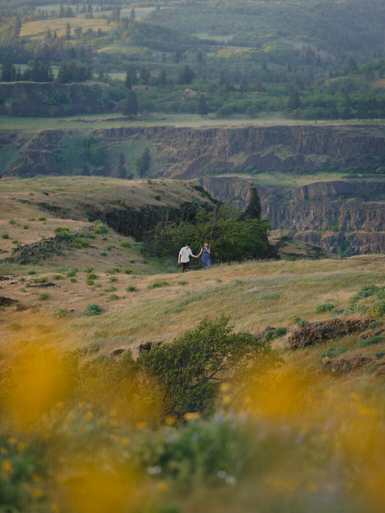 Couple strolls hand in hand through dramatic Oregon cliffs framed by wildflowers in the foreground.