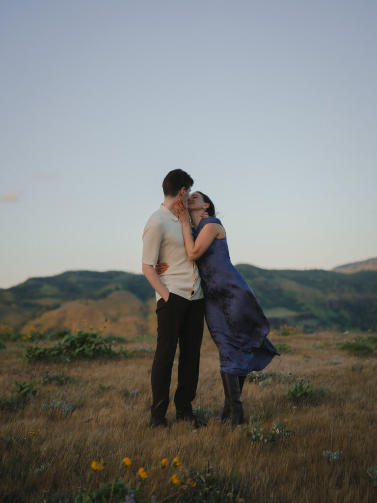 She kisses his cheek under a moody blue sky, standing close on a windswept plateau after sunset.