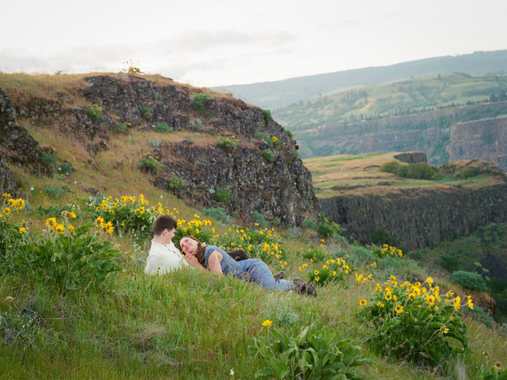 Couple lounging together on a green hillside above dramatic cliffs.